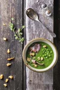 Cream soup of green peas in ceramic bowl with fried bacon and croutons. Over old wooden table with vintage spoon, salt cellar and herbs. Top view. Rustic style.